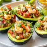 Six stuffed avocado halves filled with colorful shrimp ceviche mixture arranged on a white serving platter, garnished with fresh cilantro leaves and accompanied by lime wedges and tortilla chips