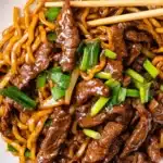 Close-up of Mongolian beef noodles coated in glossy brown sauce, garnished with green scallions and served with chopsticks on a white plate.