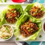 Flat lay of orange beef lettuce wraps on a serving tray with side bowls of brown rice, shredded carrots, and orange marmalade sauce for a complete meal presentation