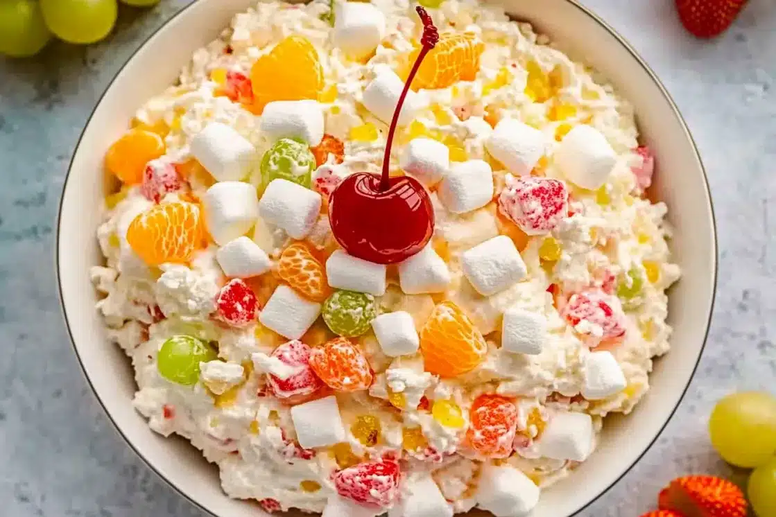 Top-down view of ambrosia salad showing the full mix of mini marshmallows, mandarin oranges, green grapes, strawberry pieces, and a central maraschino cherry garnishRéessayerClaude peut faire des erreurs. Assurez-vous de vérifier ses réponses.