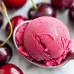 Single scoop of vibrant pink cherry sorbet on ornate silver spoon with fresh cherries in background