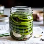 Single mason jar of pickled garlic scapes with scattered peppercorns on white cloth
