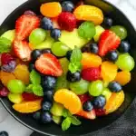 Dairy free fruit salad served in a black bowl featuring mixed berries, grapes, mandarin oranges, and fresh mint, photographed from above on marble surface