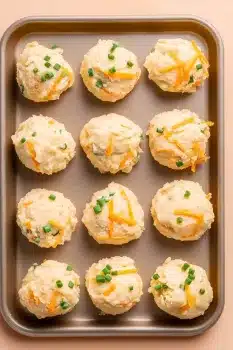 A tray of scooped mashed potato balls with visible cheddar and chives, prepped for rolling or frying.