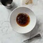 Top-down shot of a white speckled bowl containing sesame-studded soy marinade, styled on a marble background with kitchen utensils nearby
