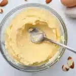 Overhead shot of frangipane cream in a glass bowl with spoon and scattered almonds on a marble surface.