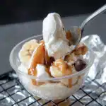 Spoon lifting vanilla ice cream from a banana boat sundae topped with caramel drizzle and chocolate chips in a clear glass bowl.