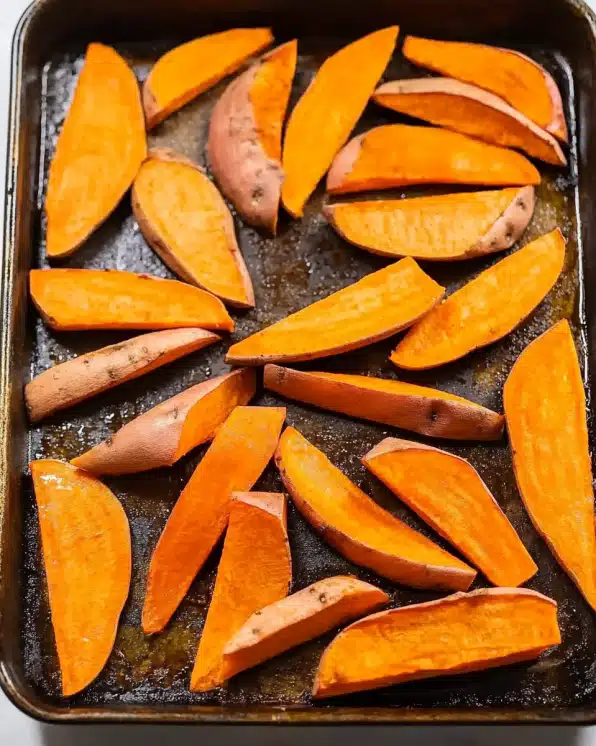 Raw sweet potato wedges arranged on a dark baking sheet before cooking, showing proper spacing and oil coating for even baking