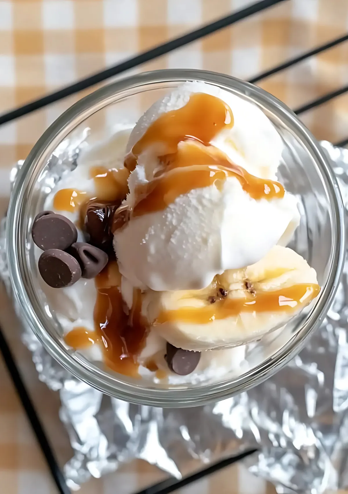 Overhead view of banana boat sundae with vanilla ice cream, chocolate chips, and caramel sauce in a glass bowl on foil.


