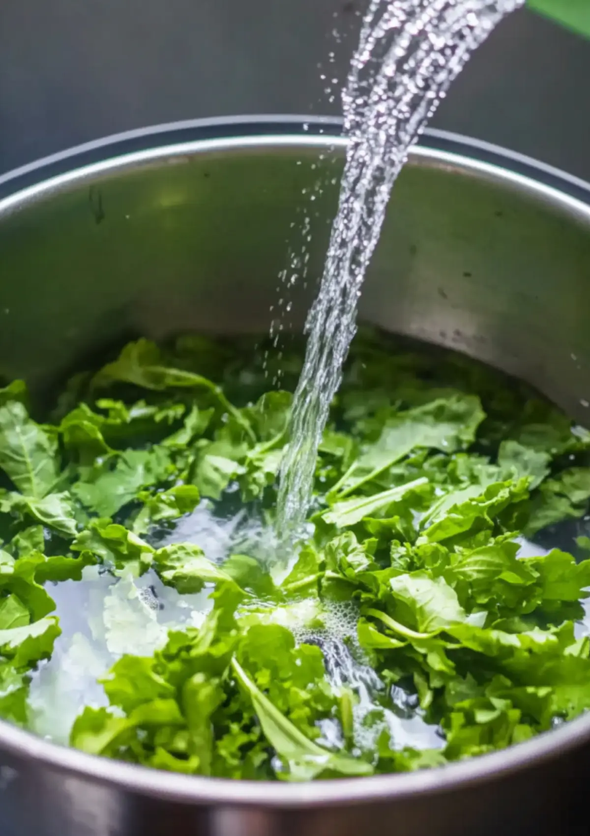 Mustard greens being rinsed in a metal bowl under running water.

