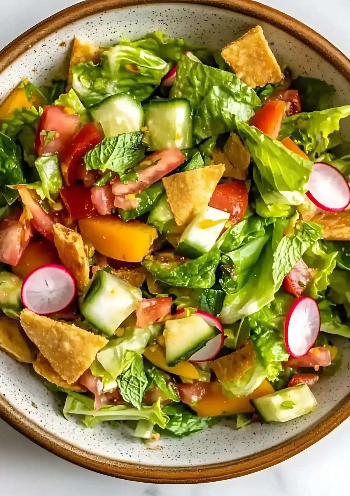 Close-up of chopped romaine, cucumbers, tomatoes, radishes, mint, and toasted pita chips in a speckled ceramic bowl