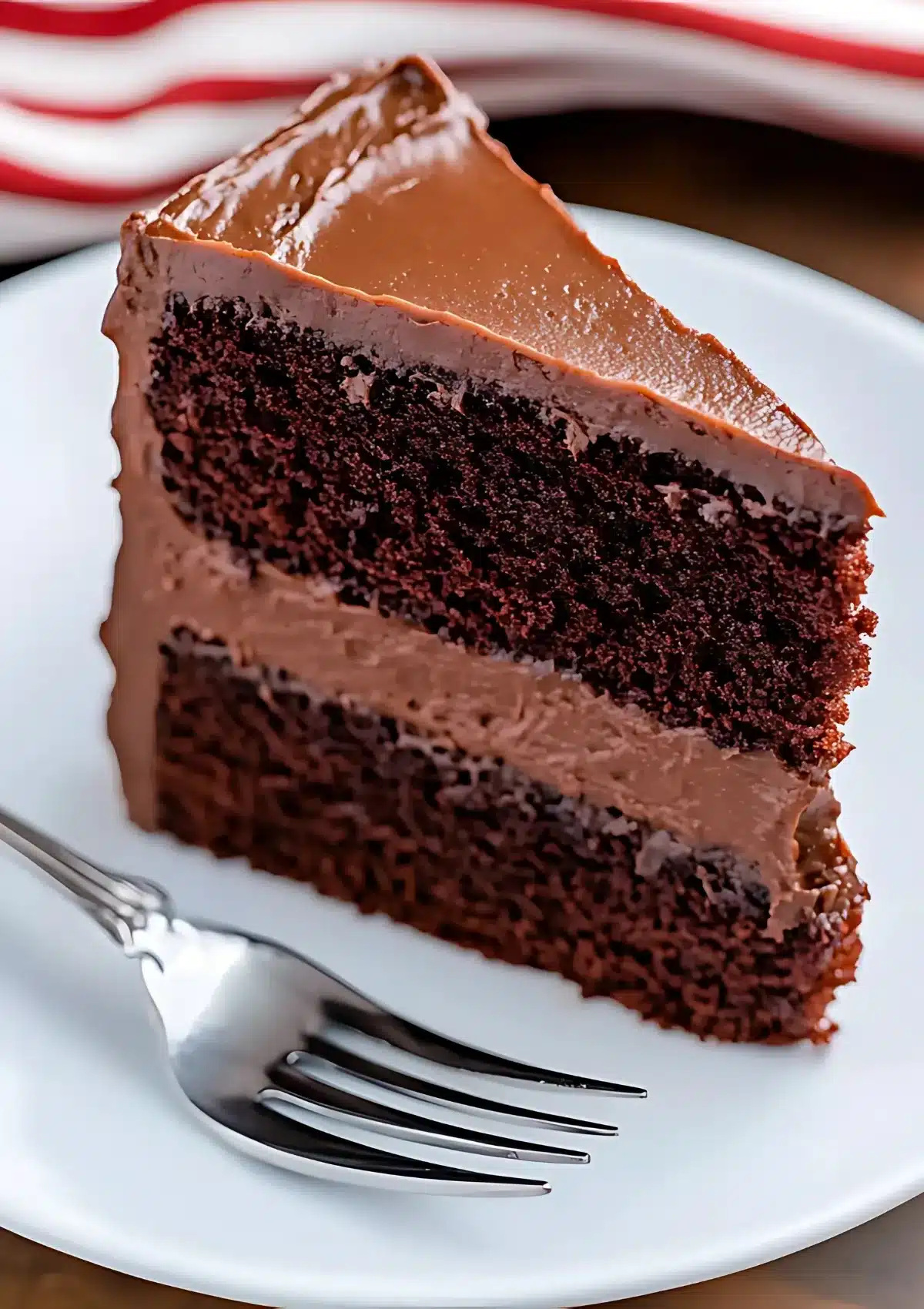 Perfect slice of moist chocolate cake with chocolate frosting on white plate with fork and red striped napkin