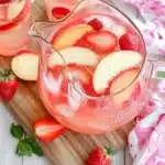 Glass pitcher of peach sangria with fresh fruit on wooden cutting board, surrounded by whole peaches and strawberries