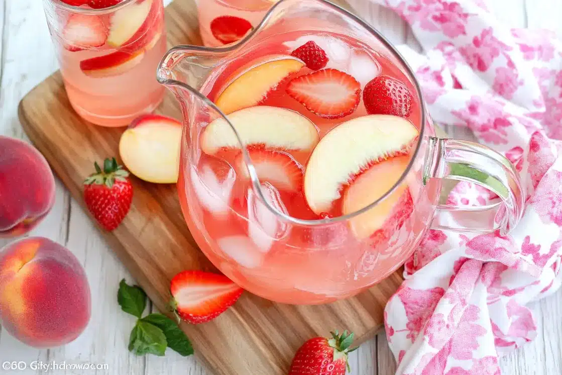 Glass pitcher of peach sangria with fresh fruit on wooden cutting board, surrounded by whole peaches and strawberries