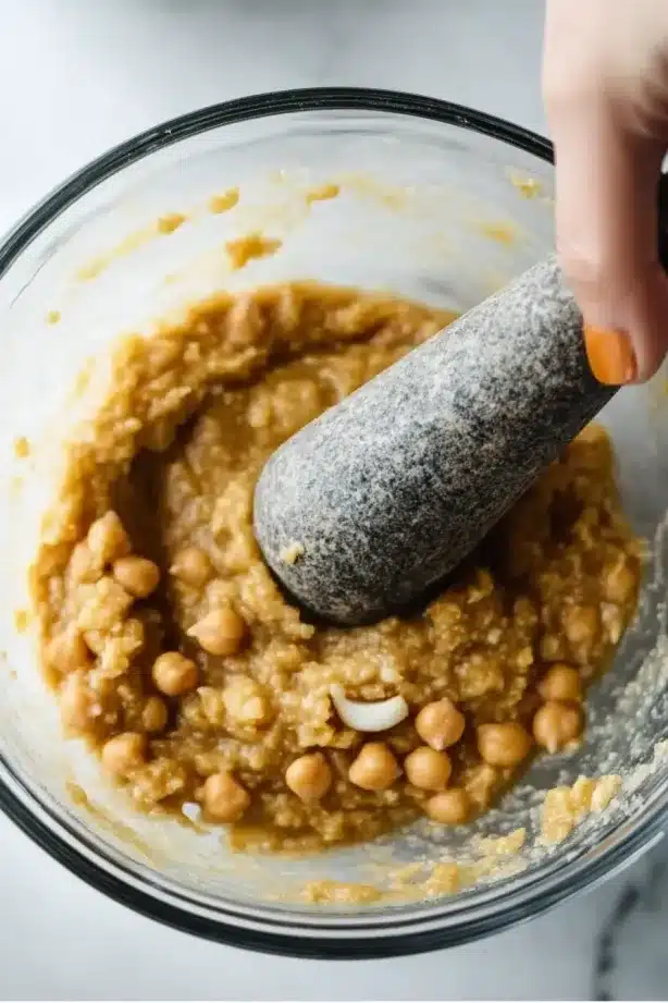Half-mashed chickpeas being processed in a bowl with a pestle, showing a thick chunky texture.