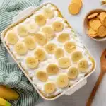 Banana pudding poke cake in rectangular baking dish with vanilla wafer cookies and fresh bananas as styling props