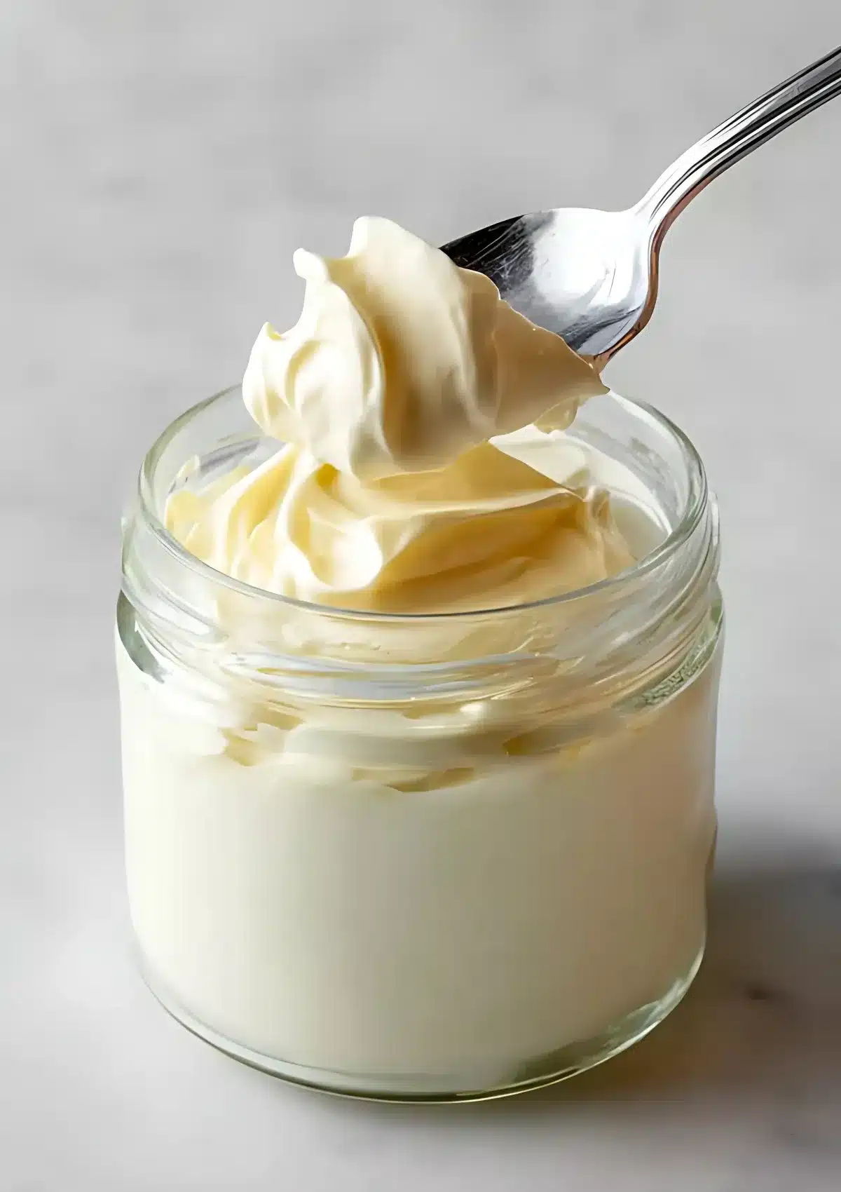 A spoon scooping velvety homemade mayonnaise from a glass jar, showing the thick and airy texture.

