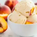Close-up of peach ice cream scoops with visible fruit bits in a minimalist white bowl, surrounded by sliced peaches.