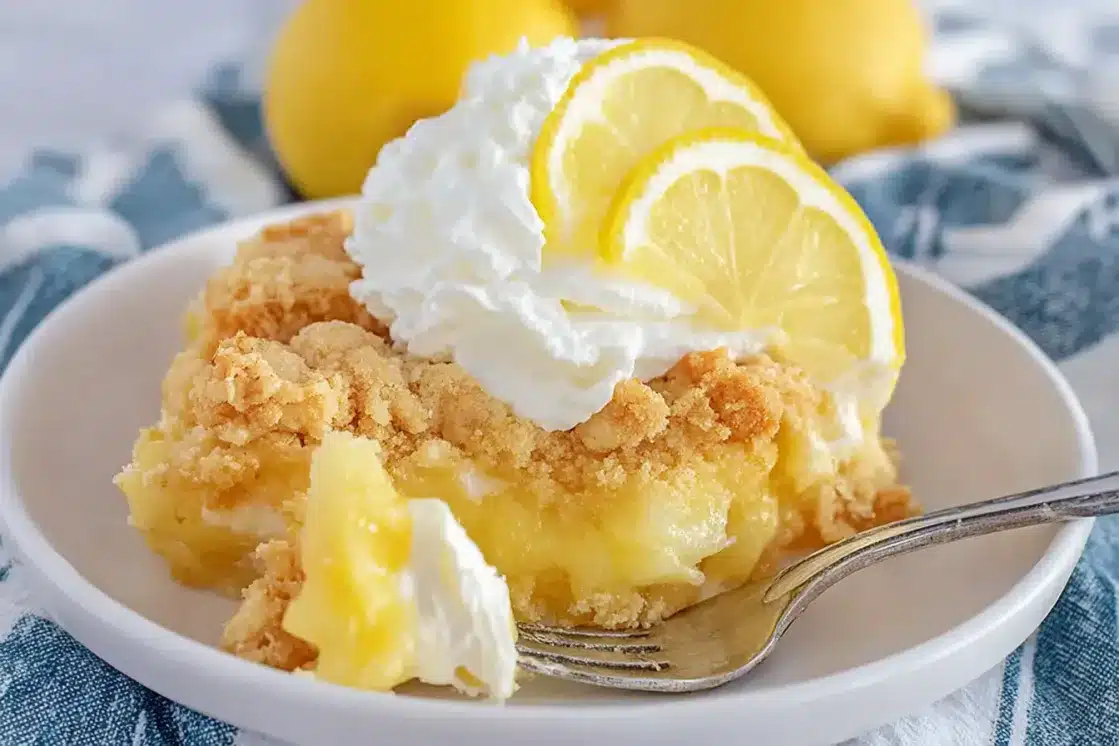 Close-up of easy lemon dump cake slice on fork showing creamy layers, golden cake crumbs, and whipped cream topping with lemon slice garnish