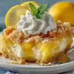 Slice of easy lemon dump cake with cream cheese on white plate, topped with whipped cream and mint garnish, showing golden crumbly texture and bright yellow lemon filling