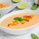 Oval bowl of chilled peach soup with peach slices and mint on marble surface with lime wedges