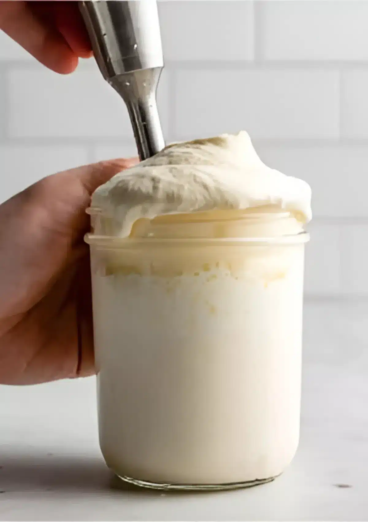 Immersion blender lifting thick homemade mayonnaise with hand holding the jar, fluffy emulsion rising to the top.

