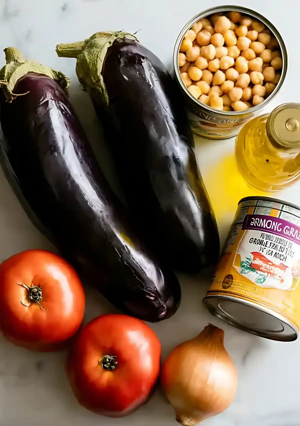 Ingredients laid out on a marble surface including eggplants, tomatoes, onion, olive oil, and canned chickpeas.

