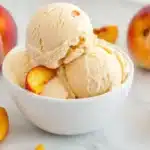 Two scoops of homemade peach ice cream in white bowl surrounded by fresh peaches and peach slices