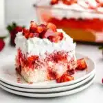 A close-up of a moist strawberry poke cake slice topped with whipped cream and fresh strawberries.