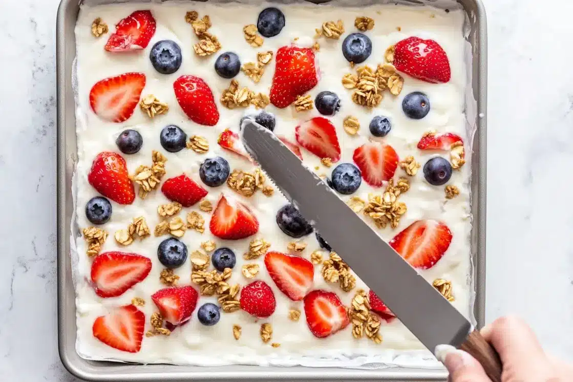 Baking sheet with yogurt base topped with fresh strawberries, blueberries and granola before freezing, hand holding knife