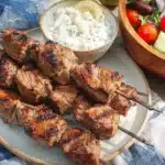 Perfectly grilled Greek lamb souvlaki skewers on white plate with creamy tzatziki sauce and traditional Greek village salad, served on blue and white striped tablecloth