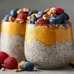 Two glasses of layered vanilla chia pudding with golden layer, topped with fresh blueberries, raspberries and crushed walnuts