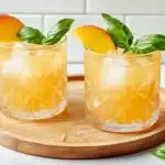 Two peach mocktails on wooden board with fresh peaches and basil leaves, white subway tile background