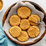 Peanut butter cookies in rustic wooden bowl with jar of peanut butter and whole peanuts on marble surface
