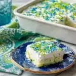 Pistachio poke cake slice on a blue speckled plate beside the full cake in a white baking dish, decorated with blue and green sprinkles on a turquoise paisley tablecloth