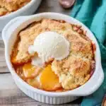 Individual peach cobbler portion in white ramekin topped with melting vanilla ice cream and copper spoons