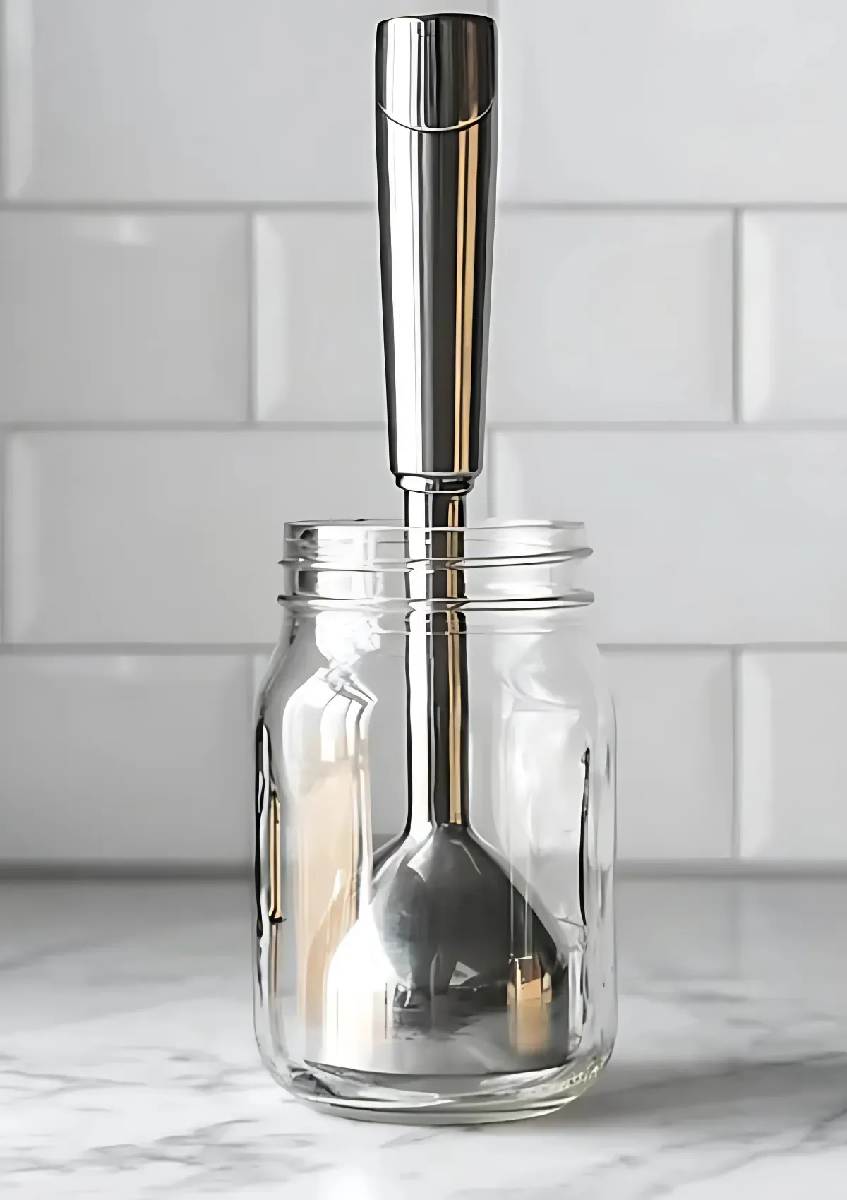 Stainless steel immersion blender placed vertically inside an empty glass jar, set on a white tiled surface.

