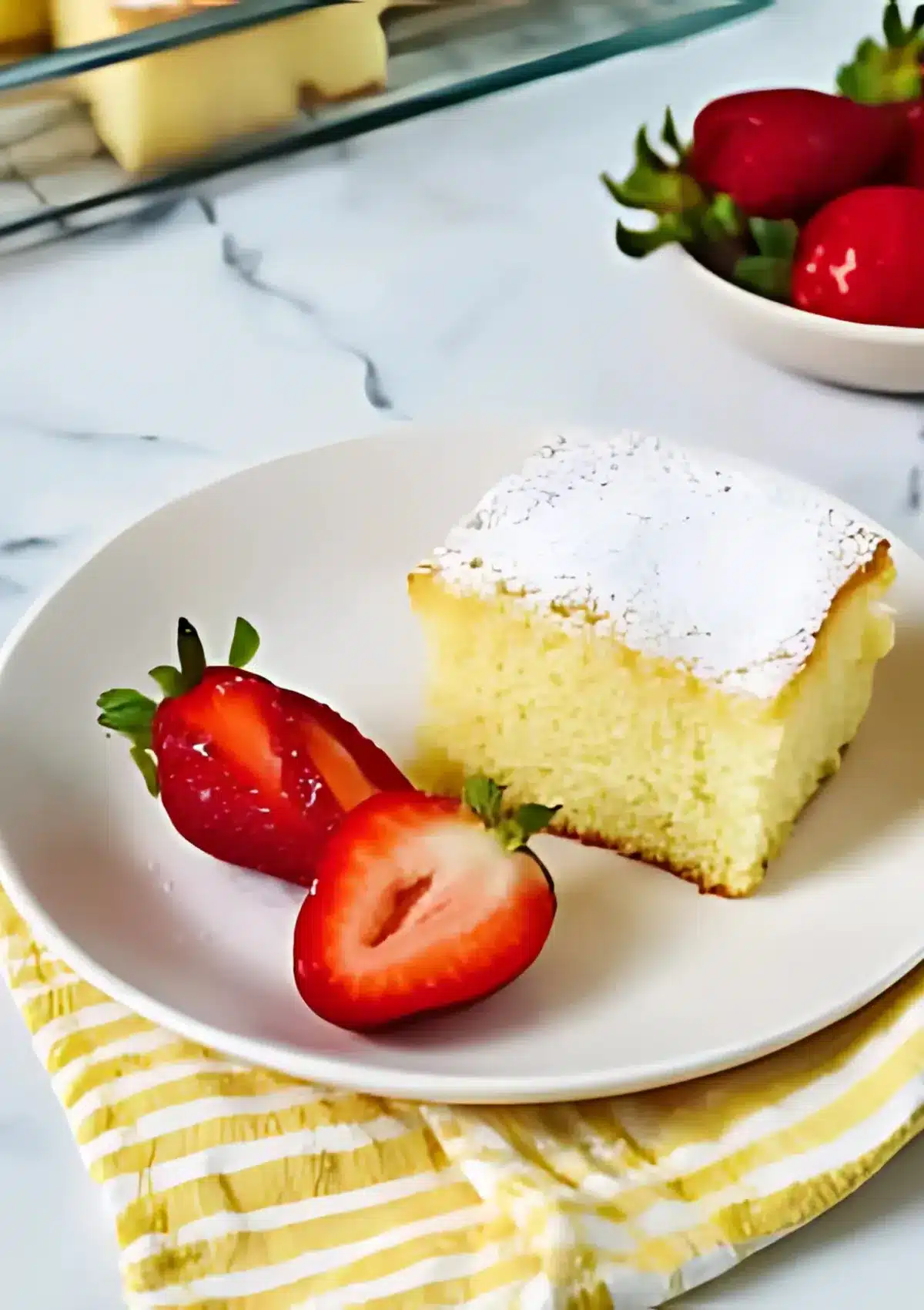 A moist square of vanilla cake topped with powdered sugar, next to halved and whole strawberries on a white plate.

