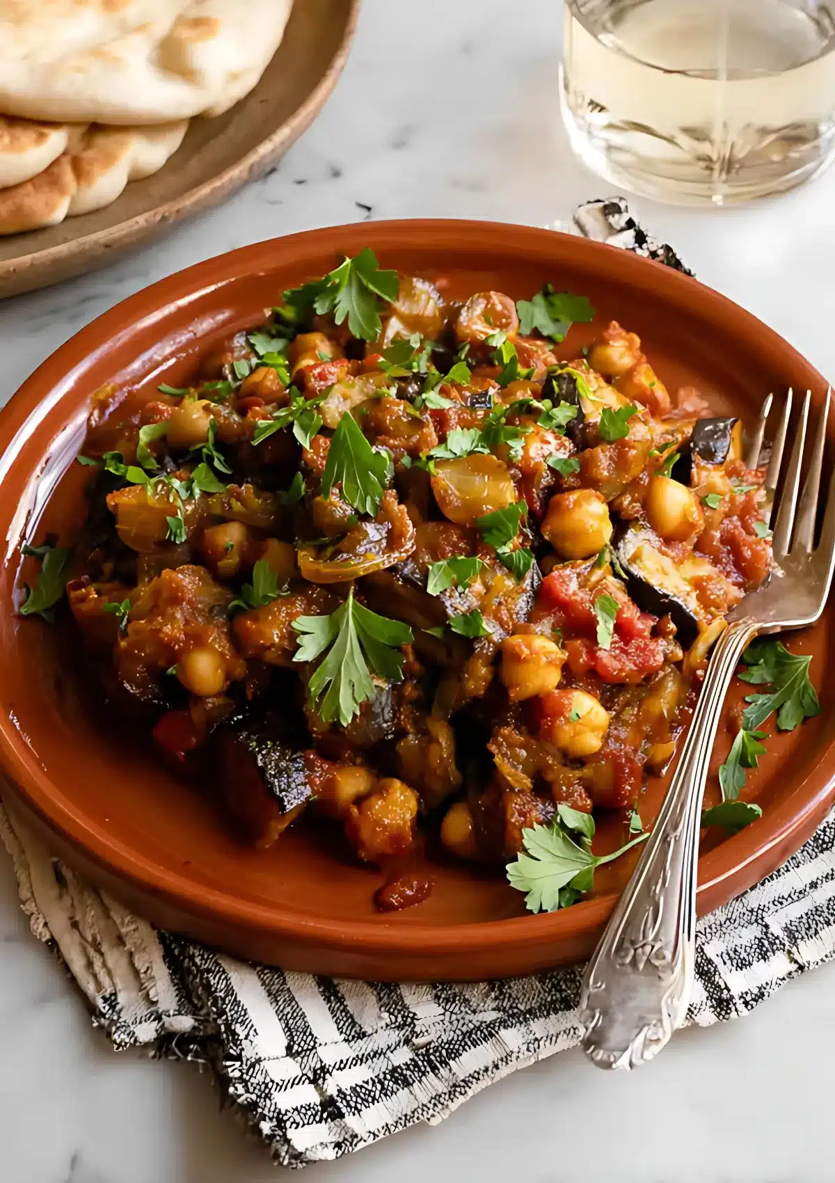 A plate of cooked Lebanese eggplant moussaka garnished with parsley, next to folded pita bread and a drink.