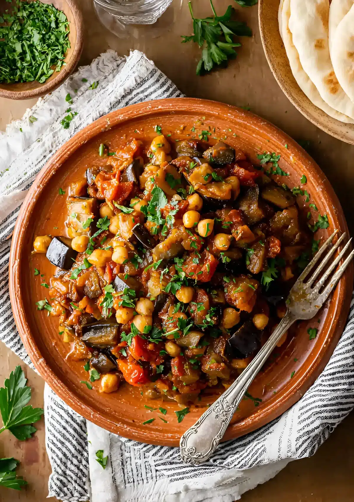 Overhead view of a plate of Lebanese eggplant moussaka served with a fork, parsley garnish, and a wooden bowl of chopped herbs on the side.

