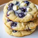 Three blueberry cheesecake cookies stacked on white plate showing golden edges and bursts of purple from blueberries