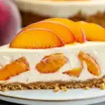 Slice of no-bake peach cheesecake on white plate showing graham cracker crust and peach pieces throughout creamy filling