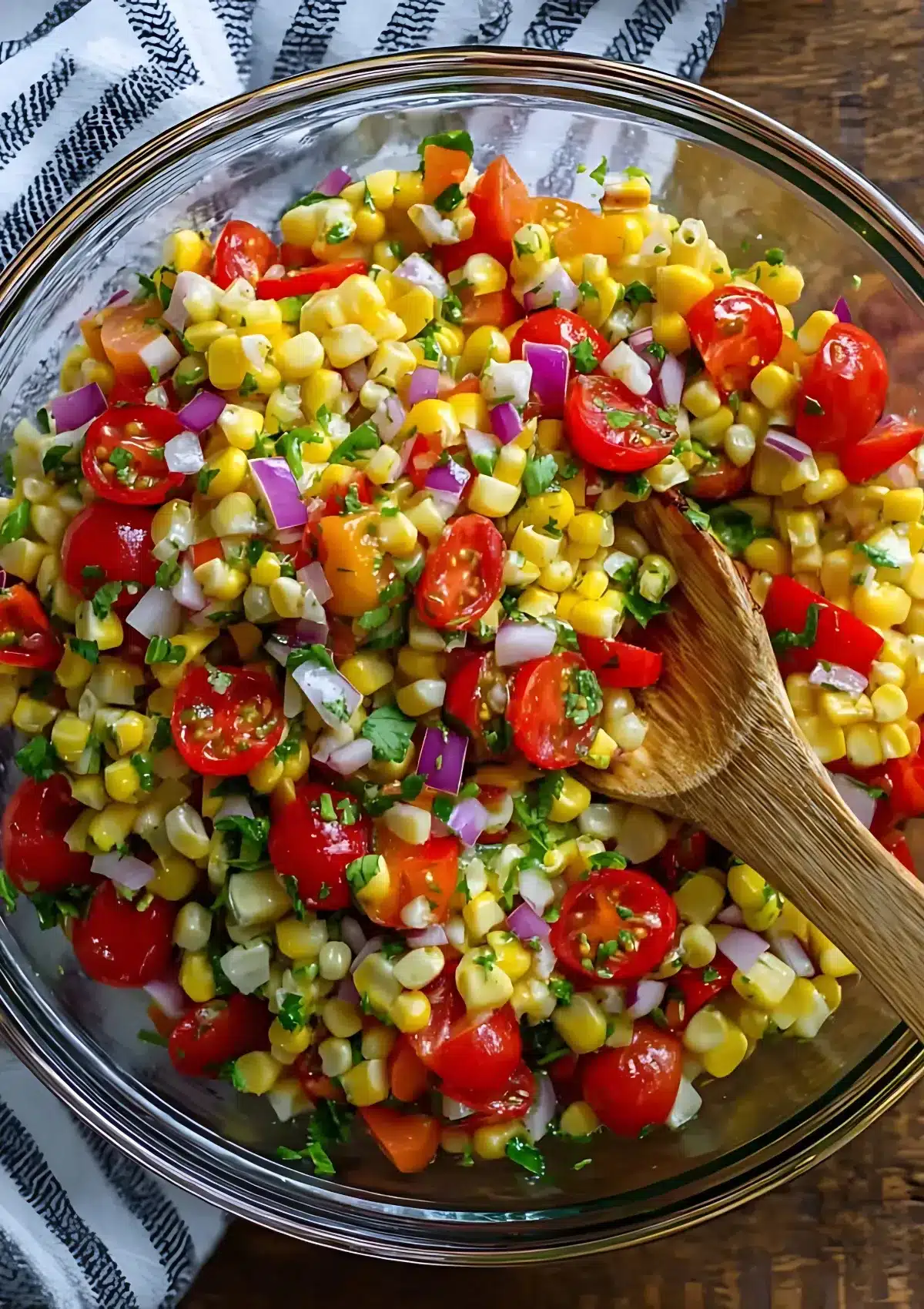 Top-down view of corn salad in a clear bowl, featuring cherry tomatoes, red onion, and cilantro, with a wooden spoon and striped towel.