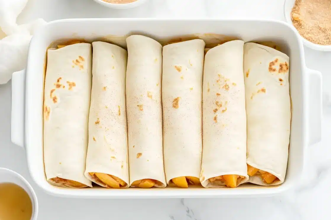 Six peach pie enchiladas rolled and arranged in a white baking dish before baking, showing the prep stage