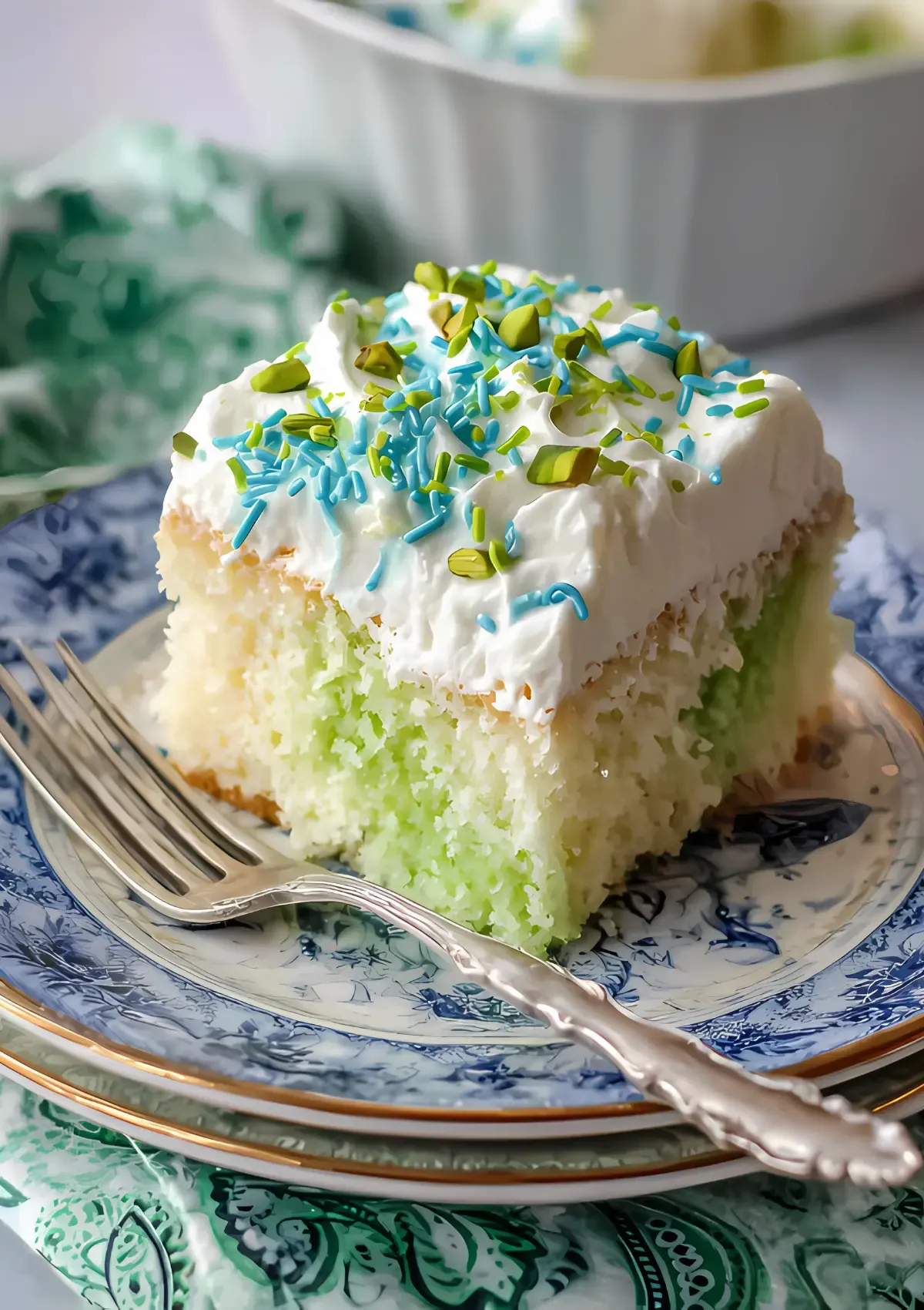 Close-up of a single slice of pistachio poke cake on an ornate plate with silver fork, highlighting the moist green cake texture and cloud-like whipped cream topping with colorful sprinkles