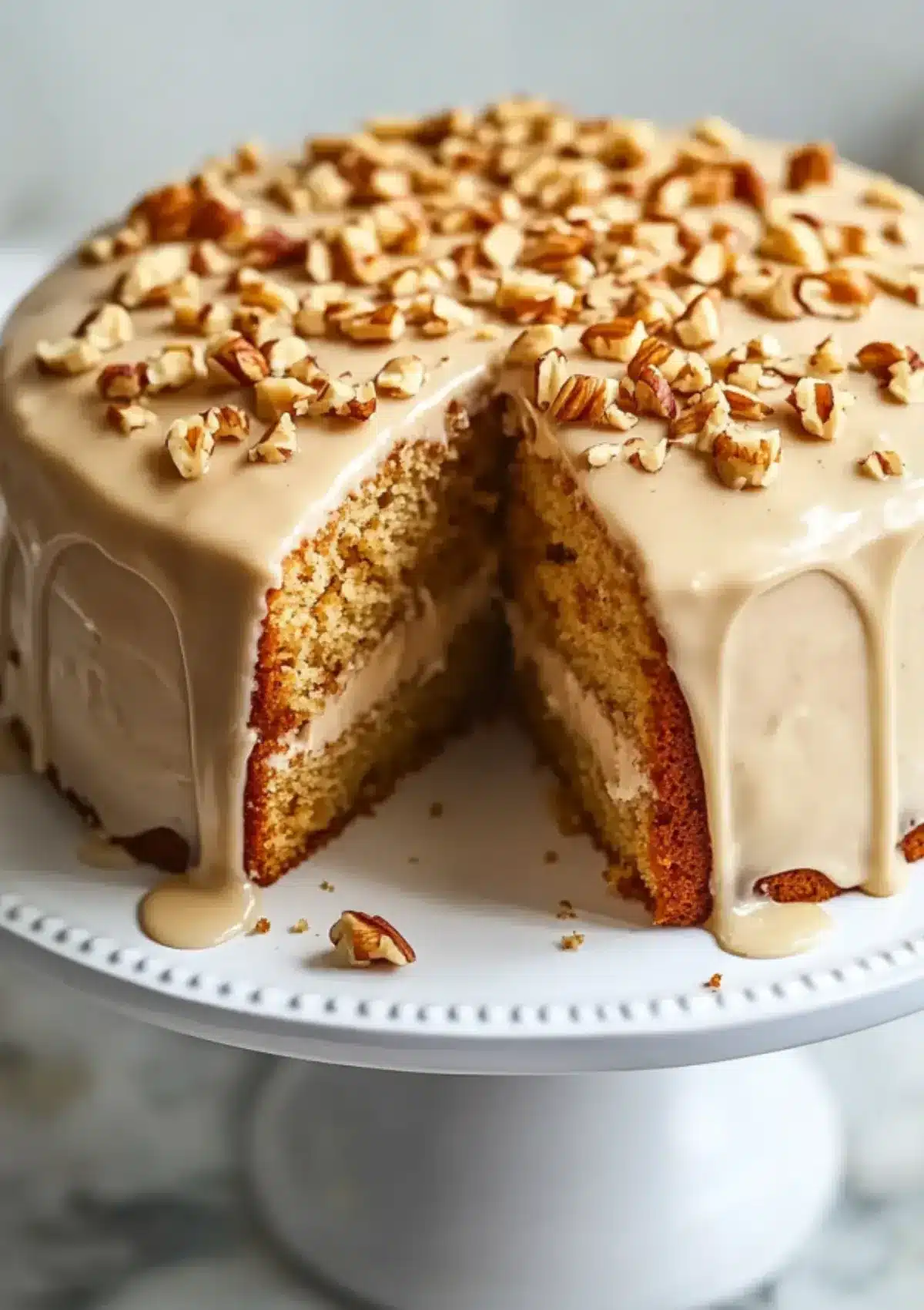Front view of a maple cream cake with thick maple frosting and chopped pecans, presented on a decorative white cake stand.

