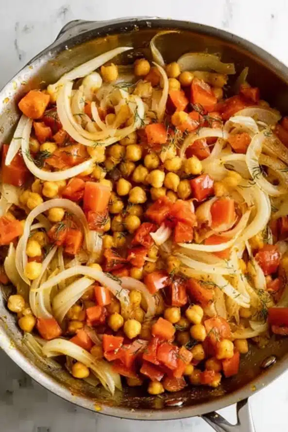 Sliced onions and chickpeas sautéed with herbs in a stainless steel pan on marble countertop.