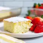 A fluffy slice of vanilla cake dusted with powdered sugar, served with fresh strawberries on a white plate.