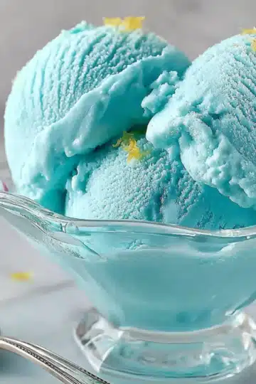 Two scoops of blue ice cream in a stemmed glass bowl topped with lemon zest, surrounded by raspberries and a silver spoon
