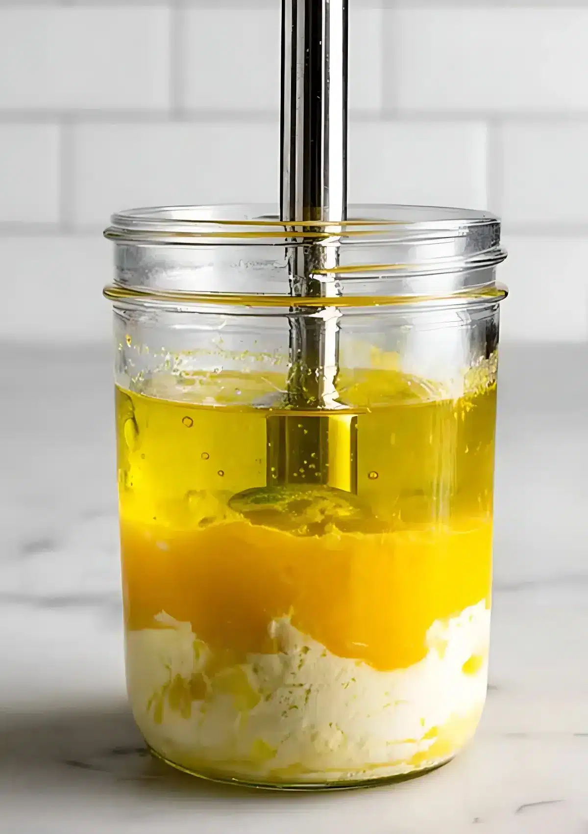 Layered egg, oil, and vinegar mixture in a glass jar before blending, with an immersion blender positioned inside.

